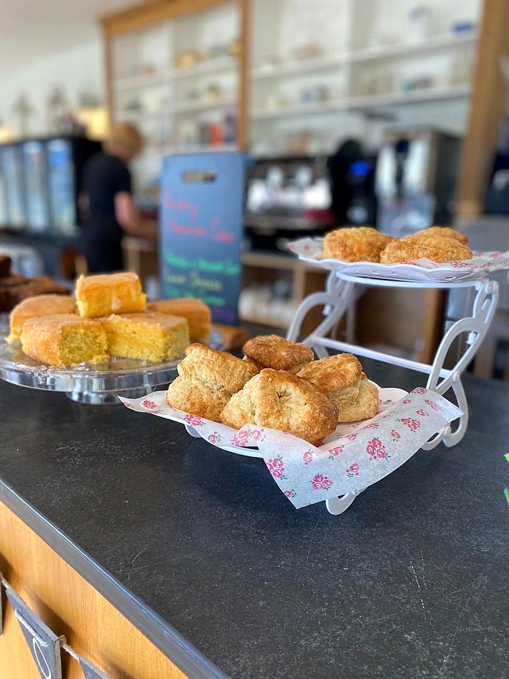 The counter: a cake stand of scones and a plate of golden sponge squares, with a chalkboard and shelves of china behind