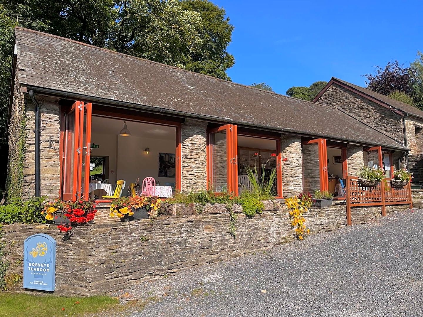 Boeveys Tearoom from the lane: a long stone building with a slate roof, red-painted posts and flower tubs, under tall trees